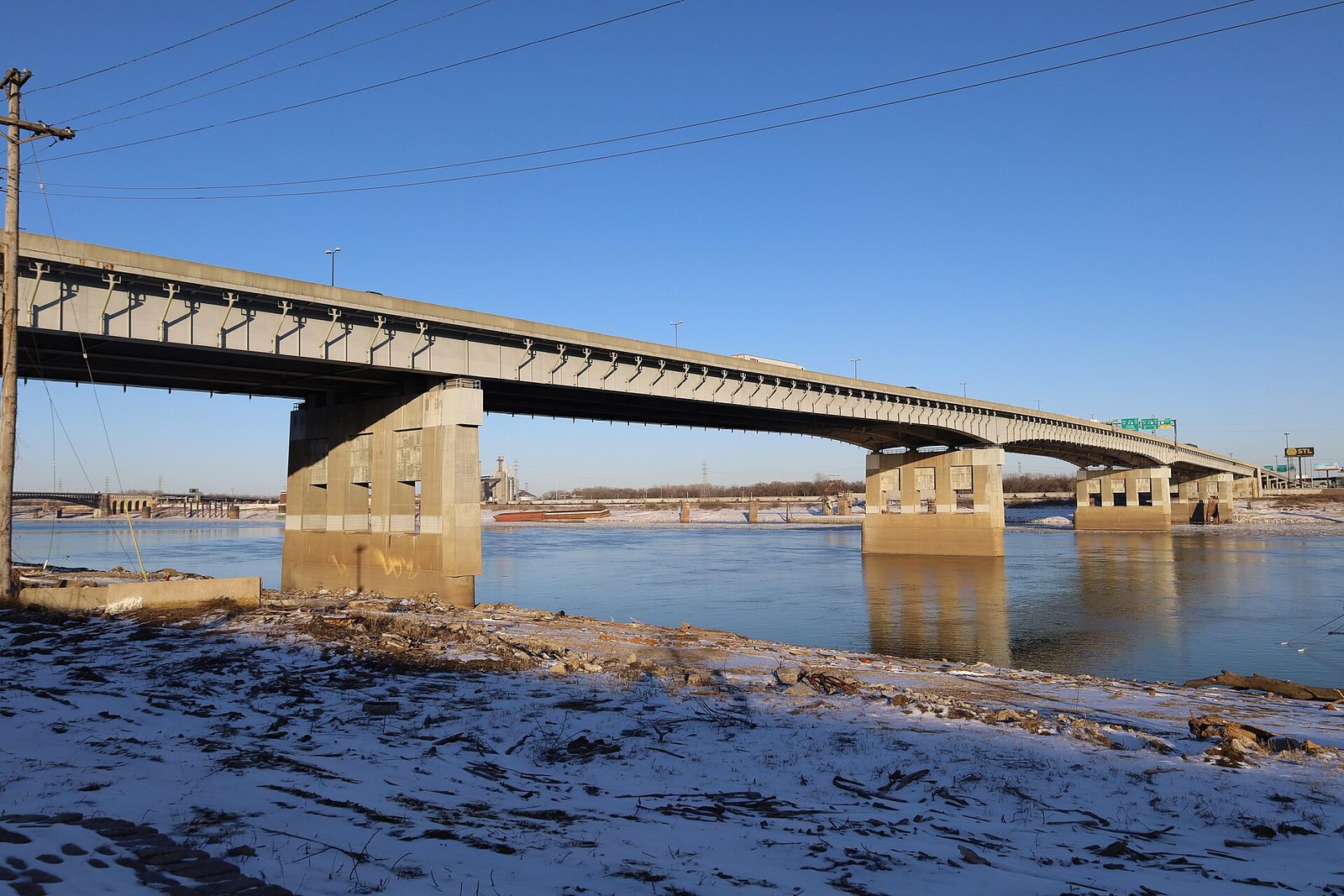 The Poplar Street Bridge over the Mississippi River on a clear winter day, concrete piers in the water and a snowy bank in the foreground